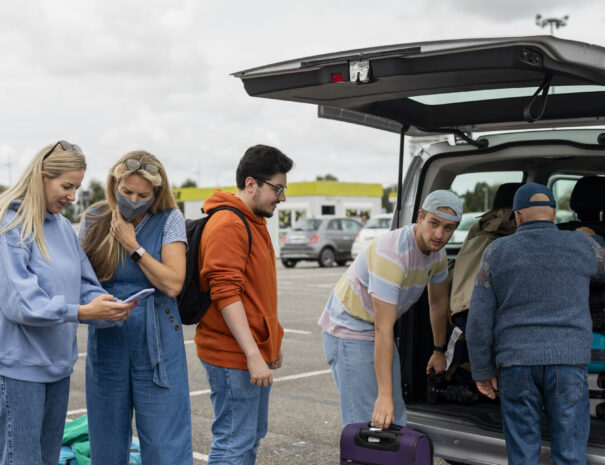 A group of friends has landed at the airport and they are in the car park loading the car trunk with their luggage.